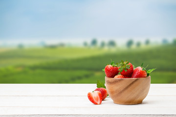 Wall Mural - Red strawberries with leaves in a wooden bowl on the wooden table and the plantations background