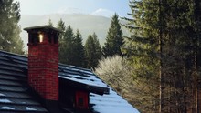 Chimney On Small Cottage House In Winter Forest, Snow On The Rooftop, Evergreen Trees In Background