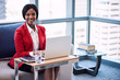 © nakophotography - Black female businesswoman smiling at the camera while seated on a couch in a business lounge with her hands on her laptop keyboard with large windows and the cityscape behind her.