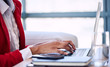 © nakophotography - Generic image of a black woman's hand busy typing on a modern notebook with her head cropped out of the image while wearing a white shirt and red blazer as well as large windows in the background.