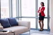 © nakophotography - Wide full body shot of business woman standing next to large windows in a business lounge, with confident body language and her arms crossed wearing formal attire
