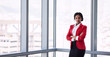 © nakophotography - formally dressed african business woman looking into the camera with her arms crossed while standing in front of large glass windows with a cityscape behind her.
