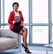 © nakophotography - square image of well dressed young black corporate woman wearing a red jacket, holding a digital tablet while sitting on the edge of the couch in a modern business lounge.