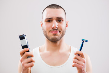 Young man is holding electric and classic razor. He is deciding which one to use to shave his beard.