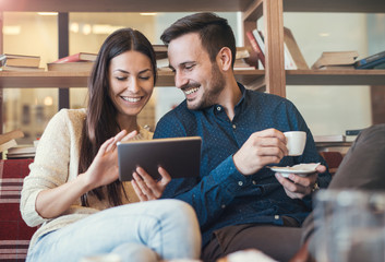 Young couple drinking coffee in cafeteria and having fun with tablet, surrounded with books on the wooden shelves