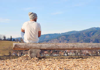 Casual young man seated on a bench in the nature looking the landscape in a sunny day with blue sky with a forest and town in the background