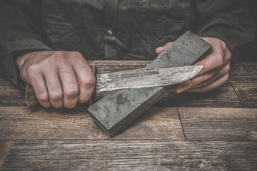 Man's hands sharpening old knife on the wooden table. Dark, vintage atmosphere.