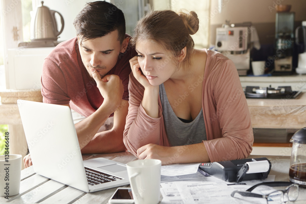 Serious man and woman sitting at kitchen table in front of open laptop computer, looking at screen with concentrated expression, focused on paying utility bills online. Family budget and finances