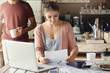 © wayhome.studio  - Happy beautiful woman reading notification from bank on prolongation of mortgage term while working through papers at kitchen table with laptop and calculator, her husband standing behind her