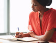 © nakophotography - Beautiful young african american writer busy making hand written notes while wearing a bright orange blouse and sitting next to large bright windows.