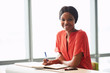 © nakophotography - Female African American writer wearing an orange blouse while looking into the camera still holding a pen in her hands that she was using to write down her notes.