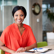 © nakophotography - Square image of happy african woman smiling while looking into camera while wearimg a colourful orange blouse and sitting in a creative business lounge.