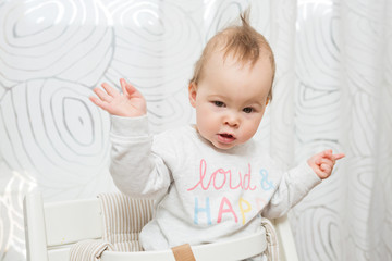 Wall Mural - Eleven months old baby girl sitting in a stool making funny faces into the camera, raising her both hands in the air