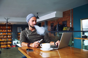 Young handsome hipster man with beard sitting in cafe with a cup of coffee, vaping and releases a cloud of vapor. Working at laptop and having a little break. With copy space.