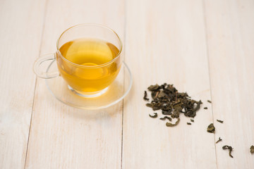 Wall Mural - Black tea in a glass cup and tea leaves in wooden spoon on white wooden background.