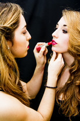 Two beautiful women Portrait where one of them paints the lips to the other over Black Background