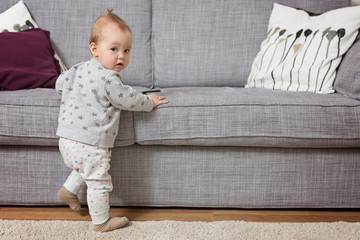 Wall Mural - Eleven months old baby girl standing by the sofa, holding it with her tiny hands