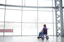 Rear View Of Disabled Woman Sitting In Wheelchair In Shopping Mall And Looking At Big Window