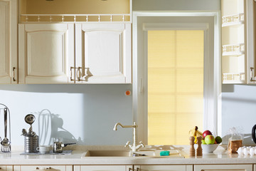 Wall Mural - Interior of lovely beige kitchen in classic style: sink with unusual tap, wooden cupboards hanging on walls, cooking utensils and products lying on stone countertop