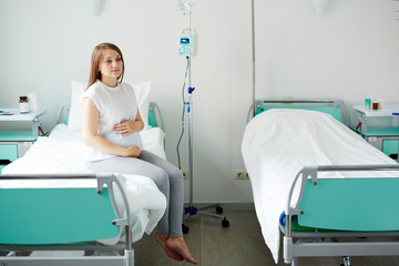 Young pregnant woman sitting on bed in comfortable ward, waiting for doctor and stroking her belly thoughtfully