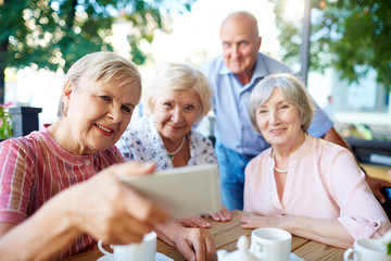 Cheerful seniors gathered together at wooden table for tea party and taking selfie with smartphone