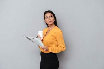 Pensive young businesswoman holding newspaper and cup of coffee