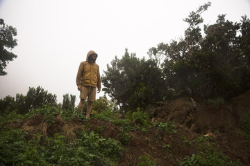 Young tourist in jacket and hood standing on green hill and looking for right way to go