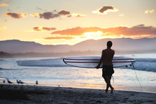 Surfer Walking On The Shore Along The Beach Lokking At The Stunning Sunset In Byron Bay, NSW, Australia.