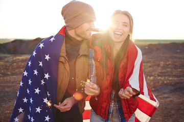 Portrait of  happy couple wearing fashionable tourist clothes laughing and  holding sparklers, both wrapped in American flag against sunlight in mountains