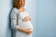 © pressmaster - Gentle blond woman expecting baby, leaning on blue wall and looking at her big belly caringly