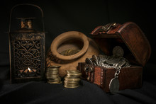 Treasure Chest, Pile And Pillar Of Coins, A Ceramic Bowl Filled With Jewelry Coins And Candle Lamp In Dark Environment 