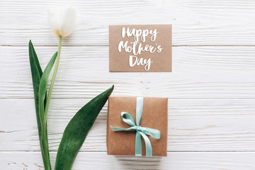 Wall Mural - happy mothers day text sign on greeting card with stylish present box and tulips on white wooden rustic background. flat lay mock up with flowers and empty paper with space for text.