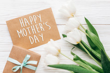 Happy Mothers Day Text Sign On Stylish Craft Present With Greeting Card And Tulips On White Wooden Rustic Background. Flat Lay With Flowers And Gift With Space For Text. Greeting Card