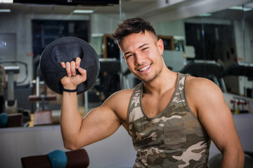 Attractive young athletic man exercising in gym, working out using kettlebell, looking at camera with a smile