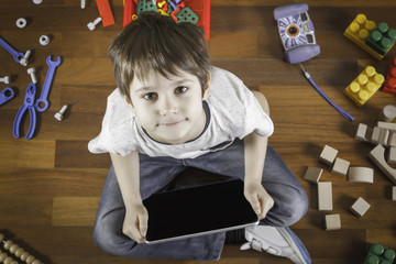 Wall Mural - Little boy playing with tablet computer. Colorful toys around him on the wooden floor. Top view