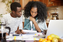 Dark-skinned Male Holding Pencil, Making Calculations On Calculator And Looking At His Worried Wife Reading Notification From Bank In Her Hands. Young African-American Couple Paying Bills Online