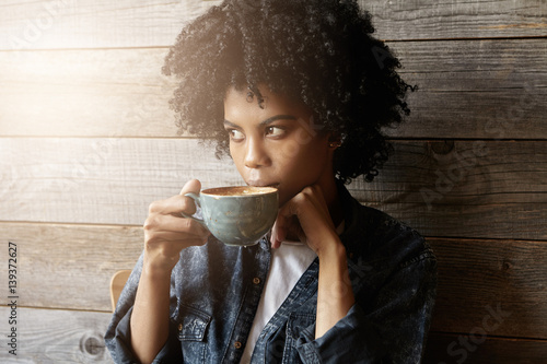Indoor shot of beautiful African-American woman with Afro hairstyle holding big mug, having fresh cappuccino and enjoying nice view outside window while relaxing alone in modern coffee shop interior