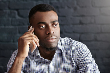 Close up shot of handsome businessman in formal wear having serious phone conversation with potential business partners, making appointment, sitting at modern cafe table against brick wall background