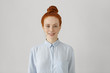 © WHstudio Leushin N - Indoor shot of attractive young redhead female with hair bun in shirt smiling happily, ready for her first day at new work. Cute student girl with freckles getting dressed before going to college