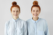 © WHstudio Leushin N - Two young redhead Caucasian females looking alike wearing same formal light-blue shirts, looking at camera, smiling happily, standing close to each other against white studio wall background