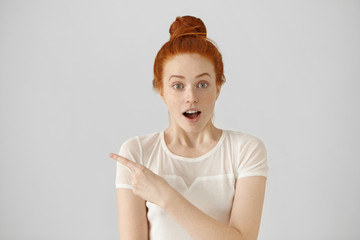 Wall Mural - Fascinated amazed young female with hair knot posing indoors at white studio wall, having surprised cheerful expression, pointing her index finger at copy space wall for your promotional information