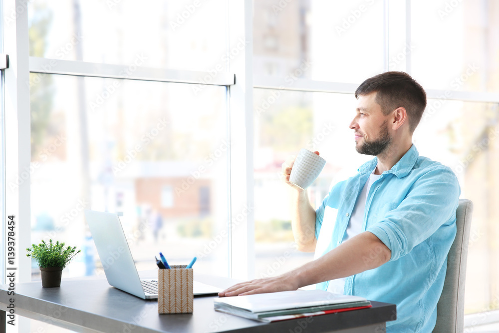 Handsome young man drinking coffee while working with laptop at home