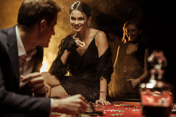 Gorgeous smiling woman with casino chip sitting on poker table and looking at man