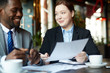 © pressmaster - Portrait of two business people meeting in modern cafe: Young professional woman smiling casually while explaining work to African-American partner and discussing documents