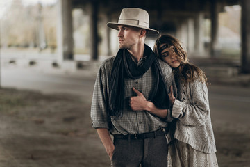 Wall Mural - stylish hipster couple gently hugging. boho woman touching arm of confident man in hat under abandoned bridge. atmospheric sensual moment. rustic fashionable look.