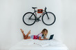 © onewithahalf - Young hipster blonde woman lays on the bed with her teddy bear while working on laptop in white room with fixed gear bicycle hanging on the wall