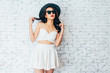 © twinsterphoto - Young happy Asian woman smiling in fashionable dress and sunglasses and summer hat over white brick wall with copy space