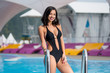 Portrait of smiling brunette girl in the black swimsuit with shapely body near the swimming pool on mountain resort with blurred background