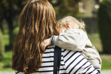 Mother carrying her toddler son for a walk in the park. Shot in the back on a warm spring day in a beautiful green garden.