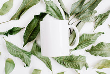 Blank Template Of White Mug And Green Leaves On White Background. Flat Lay, Top View.